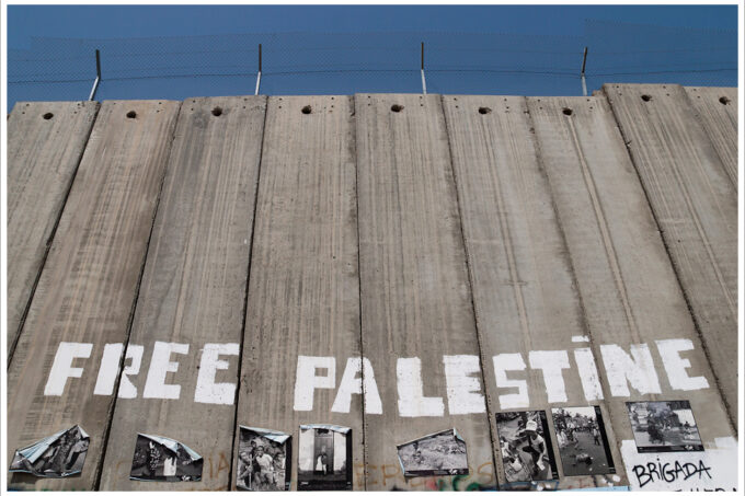Photo from below looking up at the separation wall in Bethlehem. Grey concret vertical slabs tower up with barbed wiring above. At the lower part of the wall, "Free Palestine" is painted in large white capital block letters. Below those words are black and white images pasted to the wall panels, but they are too small to break down in detail.