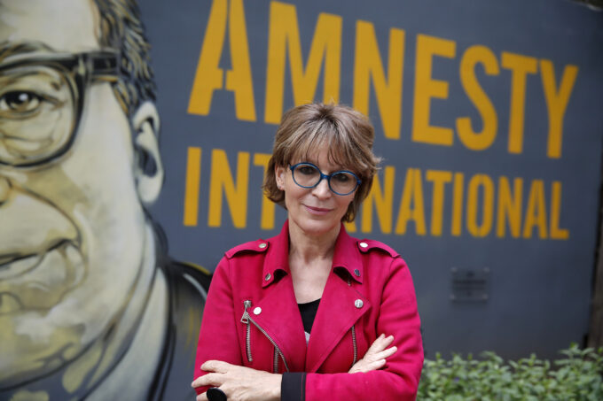 Amnesty International Secretary General Agnes Callamard poses in Paris, Tuesday, April 6, 2021. She is in the middle of the photo wearing blue round glasses and a pink jacket, crossing her arms and looking straight into the camera. She has light skin and short light brown hair with bangs. Behind her, there is a wall mural that real "Amnesty International" in yellow capital letters on a navy background. The mural also shows a cut off face of a white man with glasses and a sombre expression.