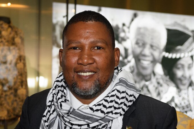 Photo of a medium-skinned Black man, the grandson of Nelson Mandela, wearing a charcoal suit jacket and a keffiyeh around his neck, with a graying goatee beard, looking into the camera and smiling. Behind him, a black and white photo on the wall of Nelson Mandela.